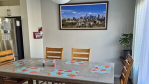 Ein Essbereich mit einem Tisch für vier Personen, Holzsesseln, einem Feuerlöscher, einem Kühlschrank und einem Stadtbild an der Wand.