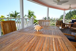 Una terraza de madera con una mesa y sillas, un ventilador de techo y vistas a palmeras y al océano.