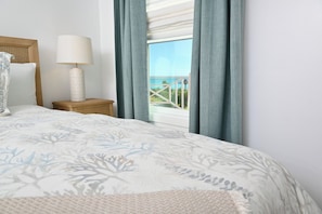 A bedroom with a bed, a nightstand, a lamp, and a window with curtains. - Luxury Beachfront House on Caribbean w/Kayaks, Paddleboards (Current)