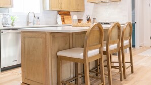 Une cuisine avec des meubles en bois, un îlot central doté de chaises, des luminaires suspendus et un lave-vaisselle visible.