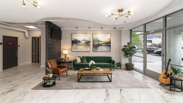 Een moderne lobby met een groene bank, een leren fauteuil en een salontafel. Er hangen twee schilderijen aan de muur, een groot raam met uitzicht, en een gitaar op een bijzettafeltje.