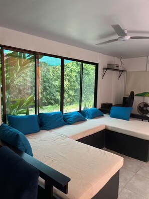 A modern room with a sectional sofa, a ceiling fan, and large windows offering a view of greenery. - Entire Downtown Home with Private Pool & A/C (Uvita)