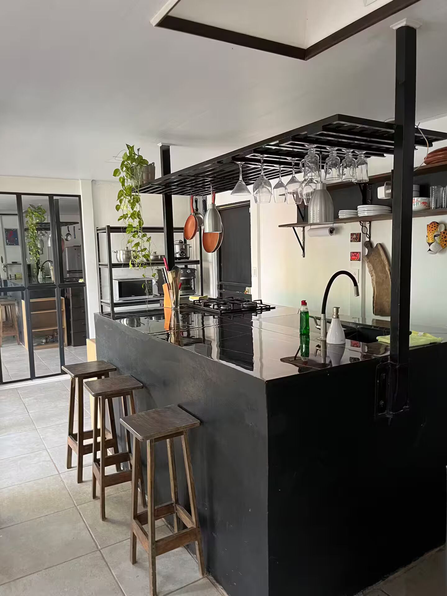 A modern kitchen with a black countertop, stainless steel appliances, hanging pots, and stools.