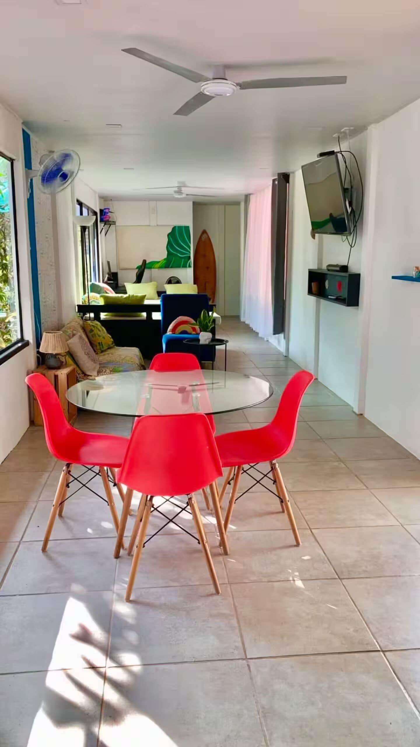 Un coin repas avec une table en verre et des chaises rouges, un ventilateur de plafond et un téléviseur fixé au mur.