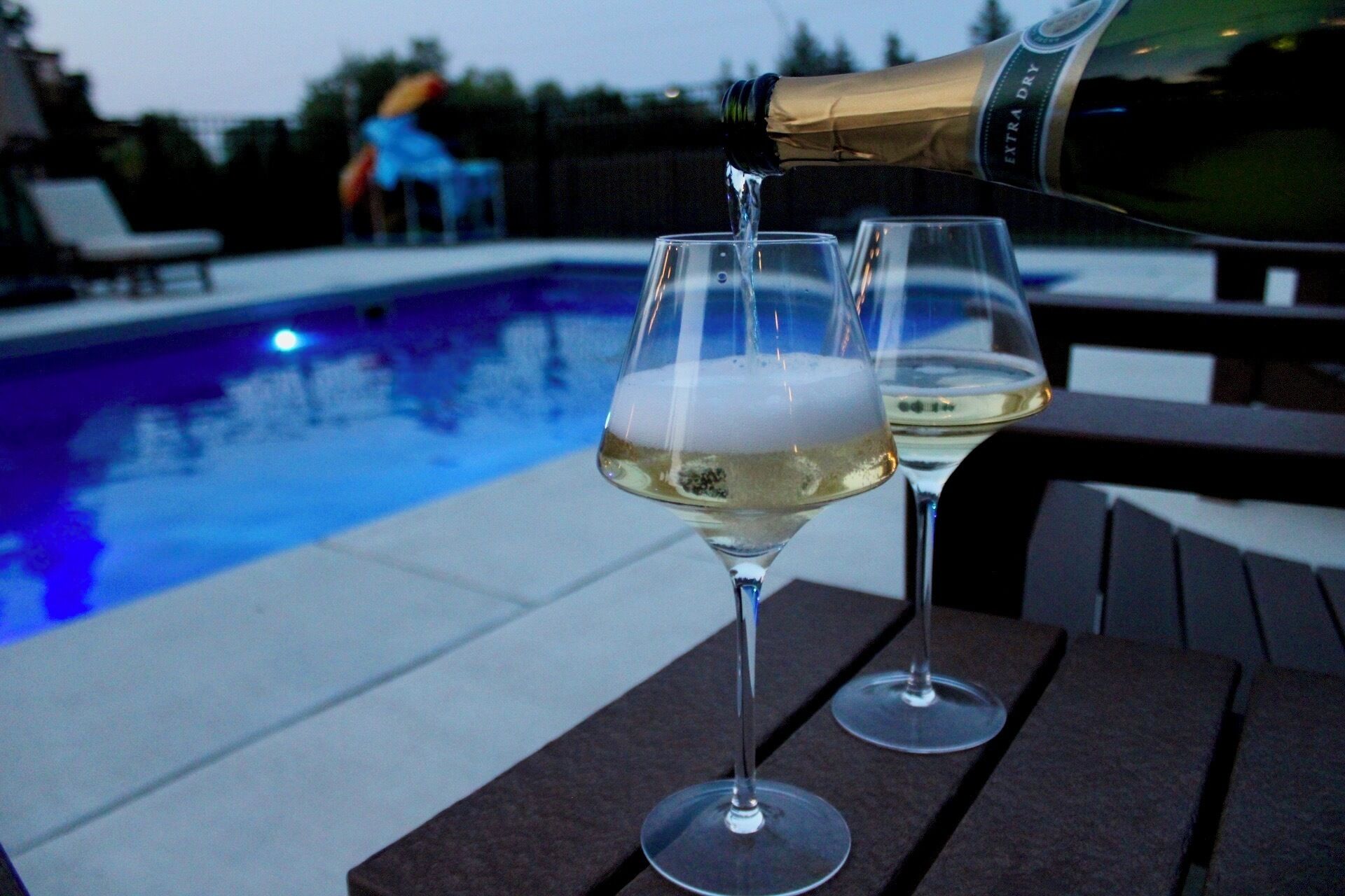 A bottle of champagne being poured into two wine glasses, with a pool in the background.