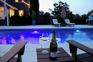 Escena junto a la piscina con una botella de vino y dos copas sobre una mesa de madera.