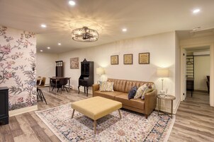 Sala de estar com sofá, banquinho e mesa de jantar com cadeiras. Há luminárias na parede e um lustre no teto.