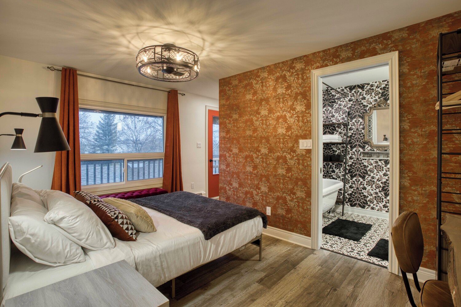 Une chambre à coucher comprenant un lit, une table de chevet, une lampe, une fenêtre avec des rideaux, une porte donnant sur une salle de bain et un plancher en bois.