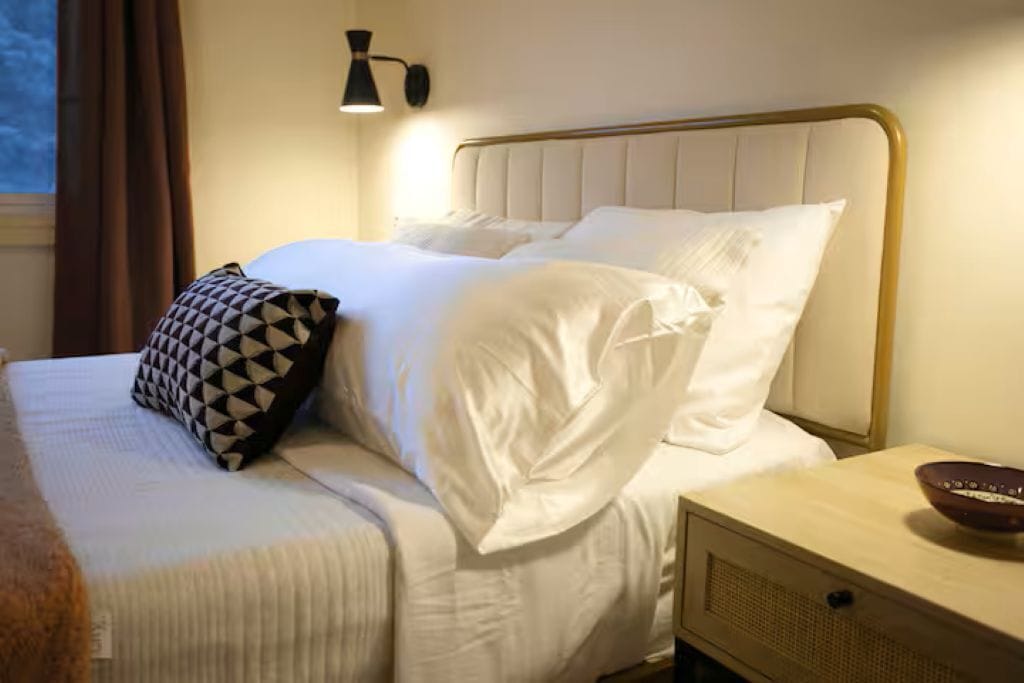 A neatly made bed with white linens and a geometric-patterned pillow, a bedside table with a bowl, and a wall-mounted lamp.