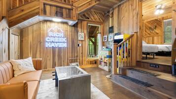 A wooden interior with a neon sign reading 'RICH CREEK HAVEN,' a sofa, a coffee table, and a staircase leading to a bedroom.
