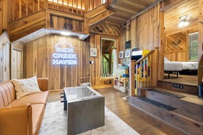 Ein Holzinnenraum mit einem Neonzeichen mit der Aufschrift 'RICH CREEK HAVEN', einem Sofa, einem Couchtisch und einer Treppe, die zu einem Schlafzimmer führt.