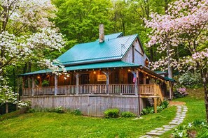 Une cabane en bois avec un toit vert, entourée d’une végétation luxuriante et d’arbres en fleurs.