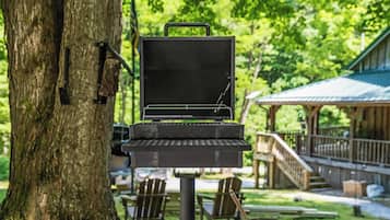 A portable grill set up against a tree in a park with outdoor seating and a covered area in the background.