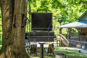 Ein tragbares Grillset vor einem Baum in einem Park, mit Sitzgelegenheiten im Freien und einem überdachten Bereich im Hintergrund.