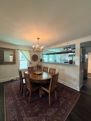A dining area with a round table, chairs, a chandelier, and a kitchen in the background. - East Nashville Bungalow, Your New Music City Homebase MidTerm Rental DOGFRIENDLY (Nashville)