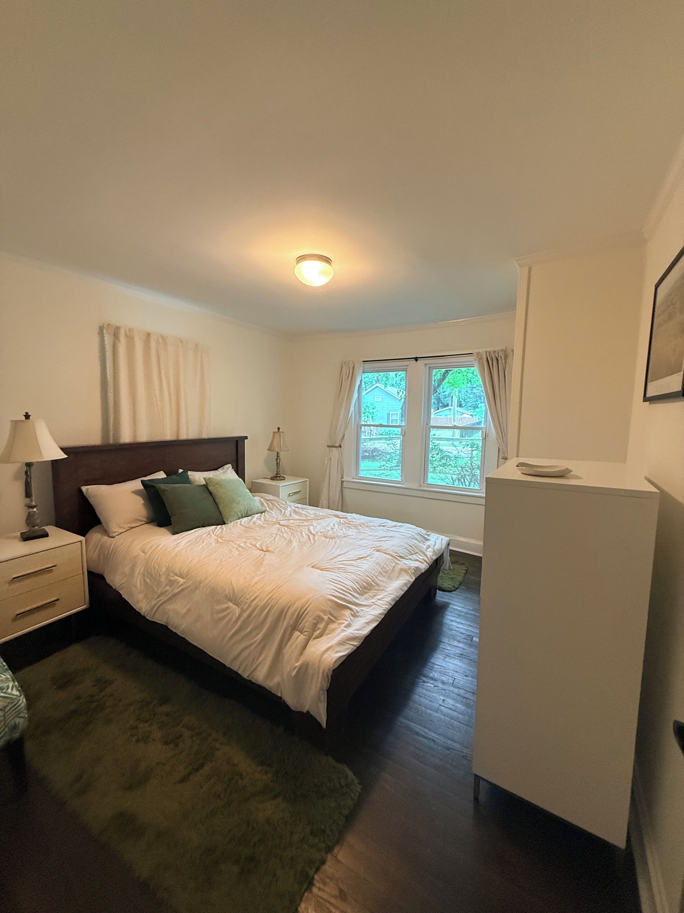 A bedroom with a bed, bedside tables, a lamp, a window with curtains, and a small cabinet.