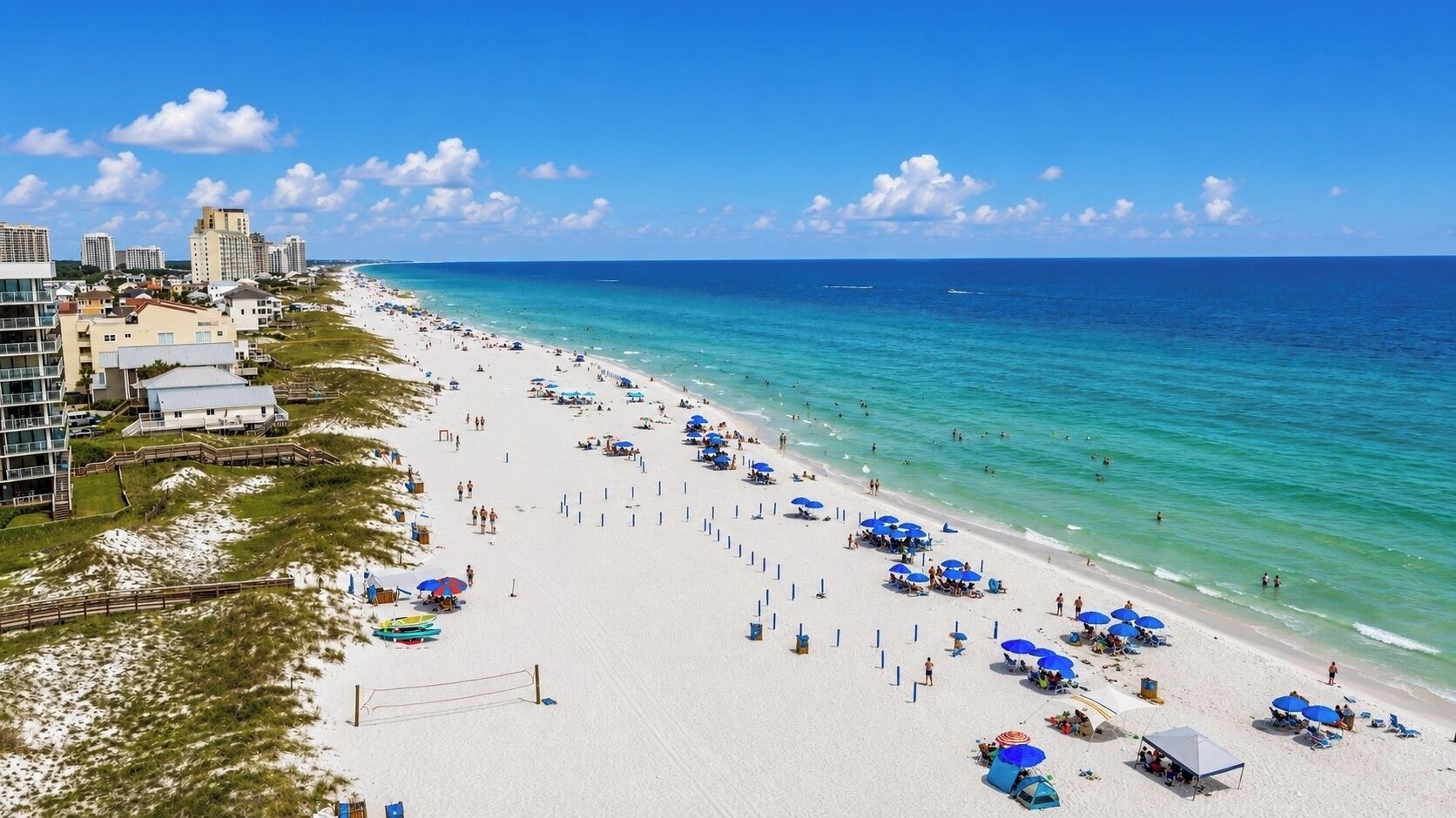 A beach with a row of umbrellas and beach chairs, a sandy shore, and buildings in the background.