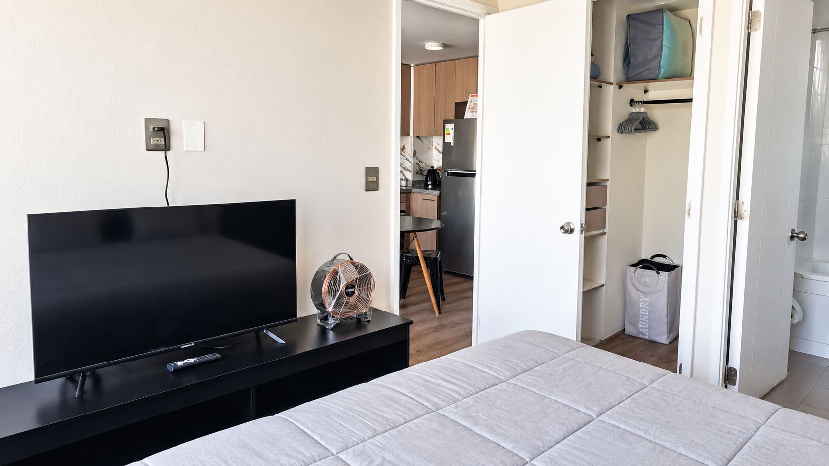 A bedroom with a TV, a fan, and a bed. There is a kitchen area visible through an open door.
