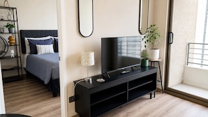 A modern bedroom with a bed, a TV on a black stand, a bedside table with a lamp, and a window with a view. - Apartment Just Steps from Line 1, the Bus Terminal, and the Train Station (Estación Central)