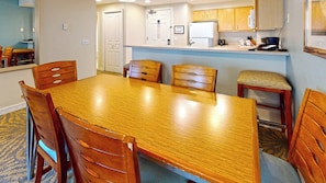 Un coin repas avec une table et des chaises en bois, une cuisine avec des placards en bois et un petit espace détente avec un tabouret rembourré.