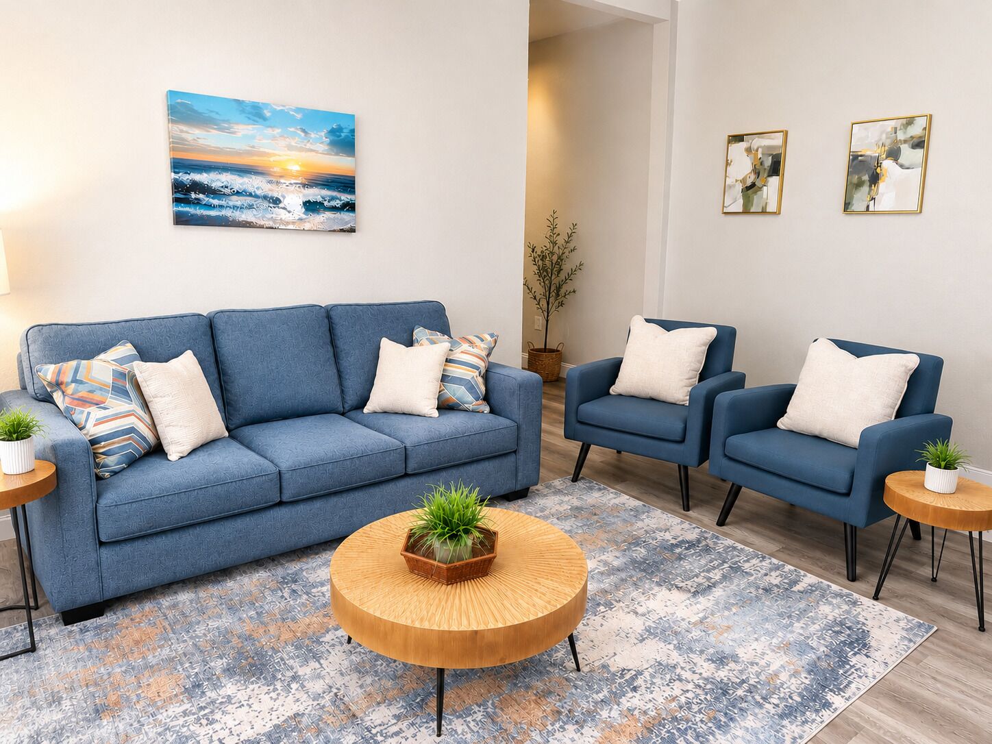 Un salon avec un canapé bleu, deux fauteuils, une table basse et un tableau représentant un coucher de soleil.