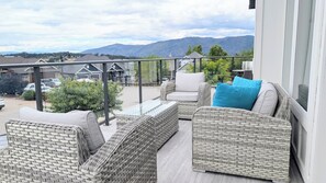 Un balcon agrémenté de meubles en osier, d’une table en verre et offrant une vue sur des maisons et des montagnes.