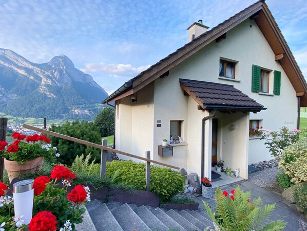 A two-story house with a sloped roof, a small porch, and a view of mountains in the background.