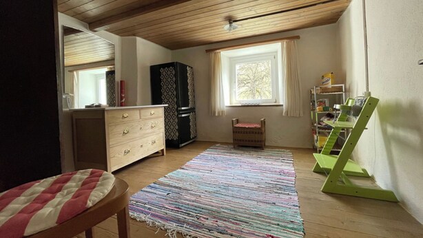 Um quarto com vigas de madeira no teto, uma janela com cortinas, um tapete estampado, uma cadeira verde e um armário.