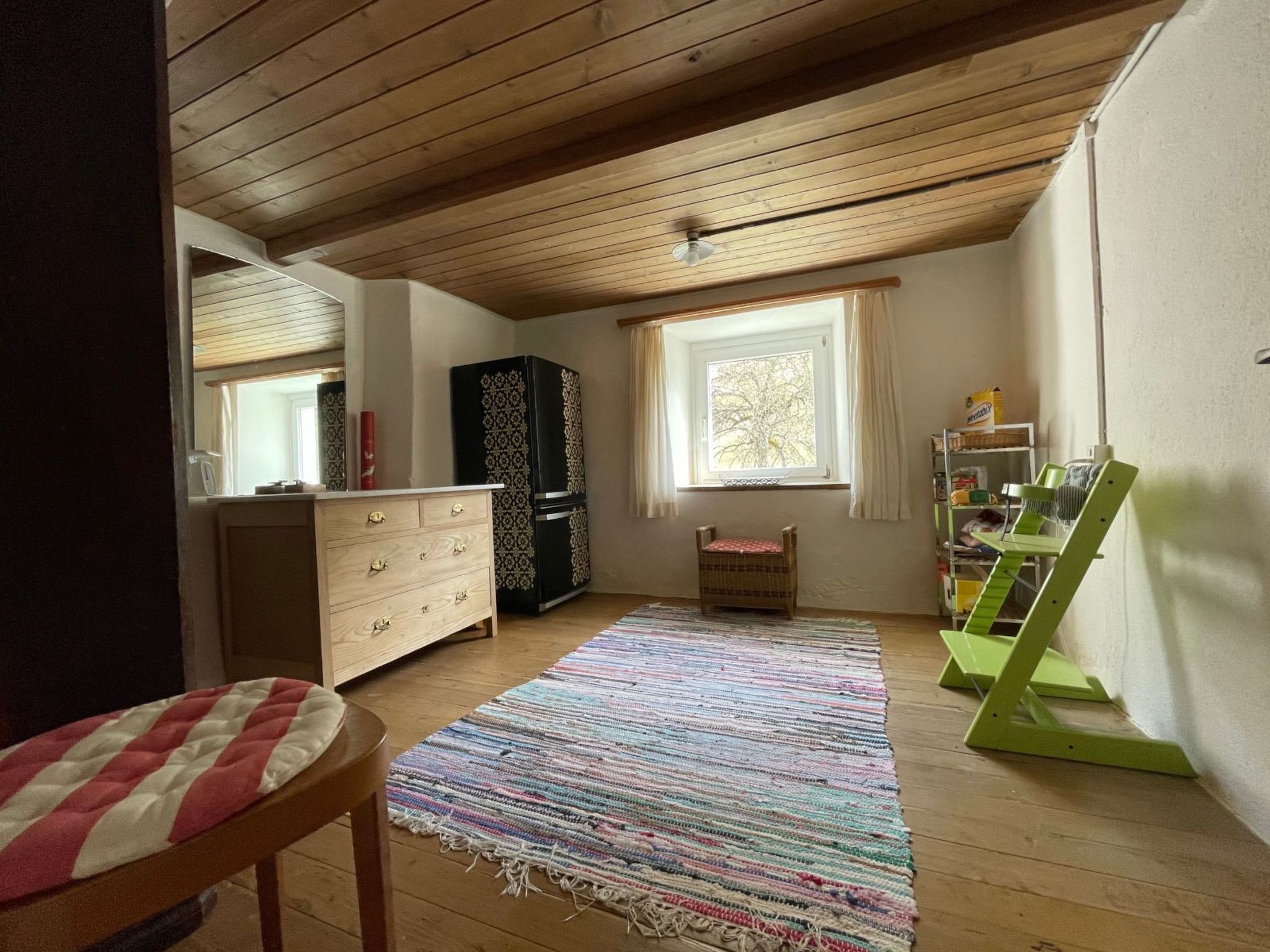 A room with a wooden ceiling, a large window, a colorful rug, a dresser, a cabinet, and a green chair.