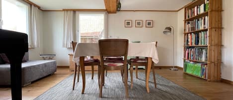 A room with a wooden floor, a dining table with chairs, a bookshelf, a sofa, and a window with curtains.