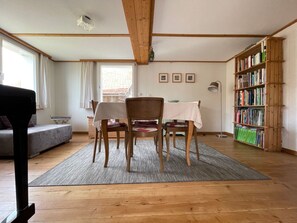 Ein Zimmer mit Holzboden, einem Esstisch mit Stühlen, einem Bücherregal, einer Couch und einem Fenster mit Vorhängen.