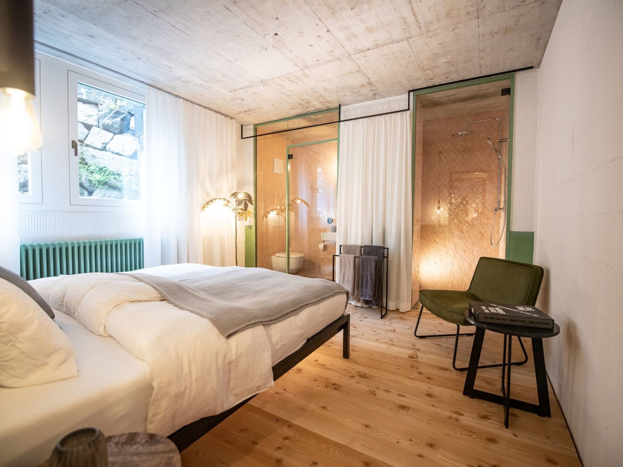 Een slaapkamer met een bed, een stoel, een radiator, een raam en een douchegedeelte.