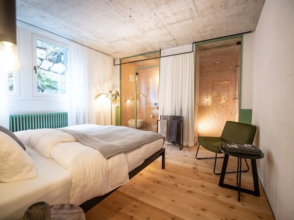 A bedroom with a bed, a chair, a radiator, a window, and a shower area.
