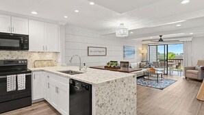 A modern kitchen with white cabinetry, a black microwave, and a dishwasher. There is a dining area with a table and chairs, and a view of the outdoors through large windows. - BayviewPalms - Sandestin 2BR Bayview + Golf Cart (Miramar Beach)