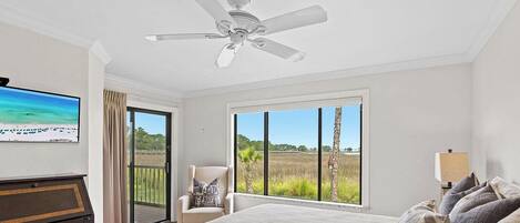 A bedroom with a bed, a chair, a desk, a ceiling fan, and large windows with a view of a beach and fields.