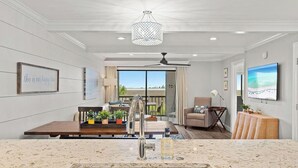A modern living room with a dining area, a ceiling fan, a TV, and a large window offering a view. - BayviewPalms - Sandestin 2BR Bayview + Golf Cart (Miramar Beach)