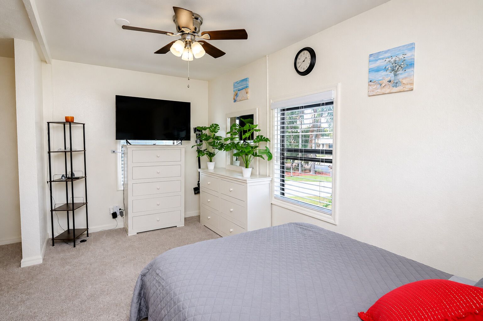 A bedroom with a bed, a dresser, a TV, a ceiling fan, a window with blinds, and a clock on the wall.