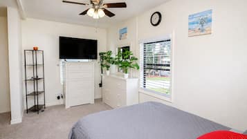 A bedroom with a bed, a dresser, a TV, a ceiling fan, a window with blinds, and a clock on the wall.
