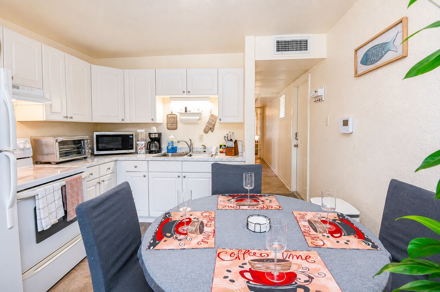A compact kitchen with white cabinets, a microwave, toaster, and a coffee maker. There is a dining table with a coffee-themed table runner and two chairs.