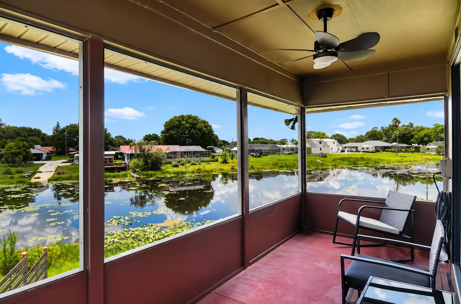 A room with large windows, a ceiling fan, and a view of a pond with houses in the background.