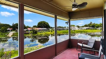 A room with large windows, a ceiling fan, and a view of a pond with houses in the background.