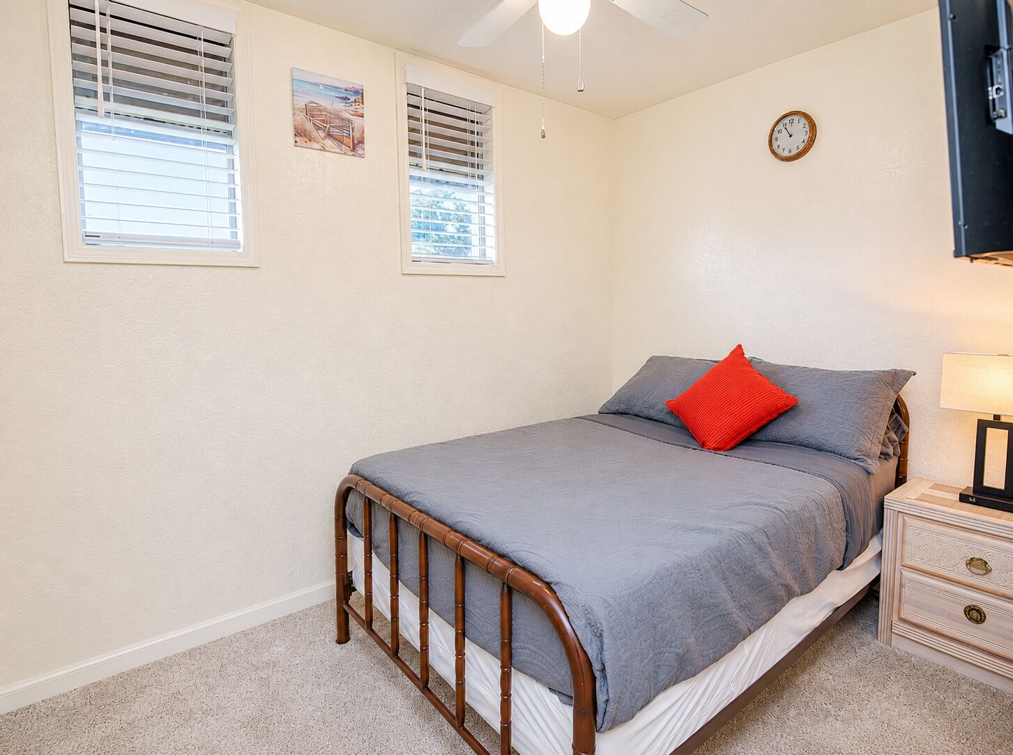 A bedroom with a bed, a nightstand, a lamp, a clock, and two windows with blinds.