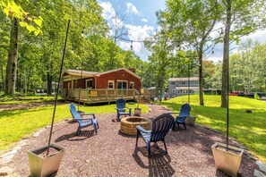 Une terrasse en bois avec des sièges d’extérieur et un foyer, entourée d’une pelouse et d’arbres.