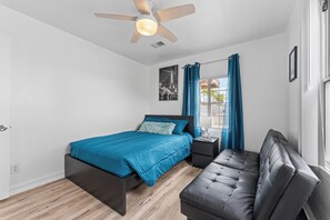 Une chambre à coucher comprenant un lit, une table de chevet, un banc, un ventilateur de plafond, une fenêtre avec des rideaux et un tableau accroché au mur.