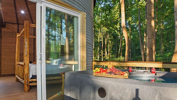 A wooden cabin with a glass door, a hot tub, and a deck overlooking a forest. - Honeymoon Suite w/ Exotic Animals + Hot Tub + Zoo (Troutman)