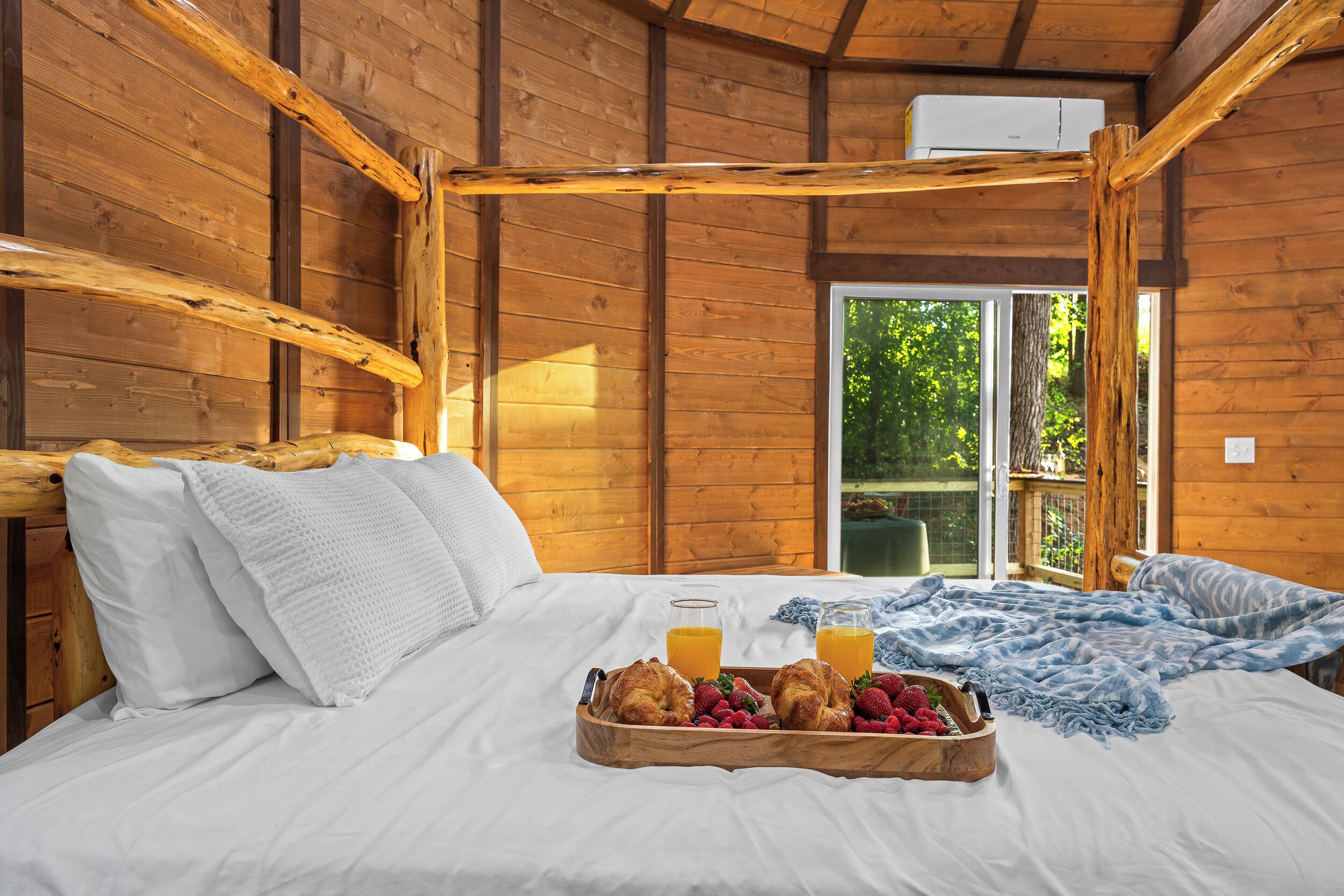 A wooden cabin room with a bed, breakfast tray, and a view of greenery outside.