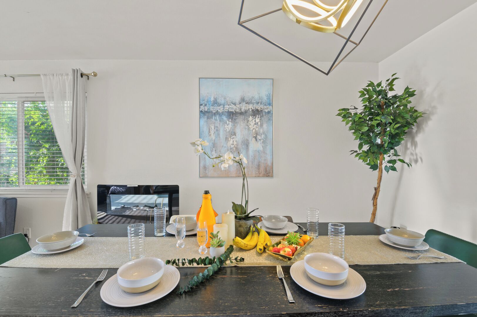 Une table à manger est dressée avec des assiettes, des verres et un saladier rempli de fruits. Un tableau orne le mur et une plante en pot se trouve sur la droite.
