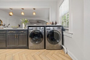 A laundry room with two front-loading washing machines, a wooden herringbone floor, and pendant lighting. - Casa Serena · Historic Spanish Home · SoSo (West Palm Beach)