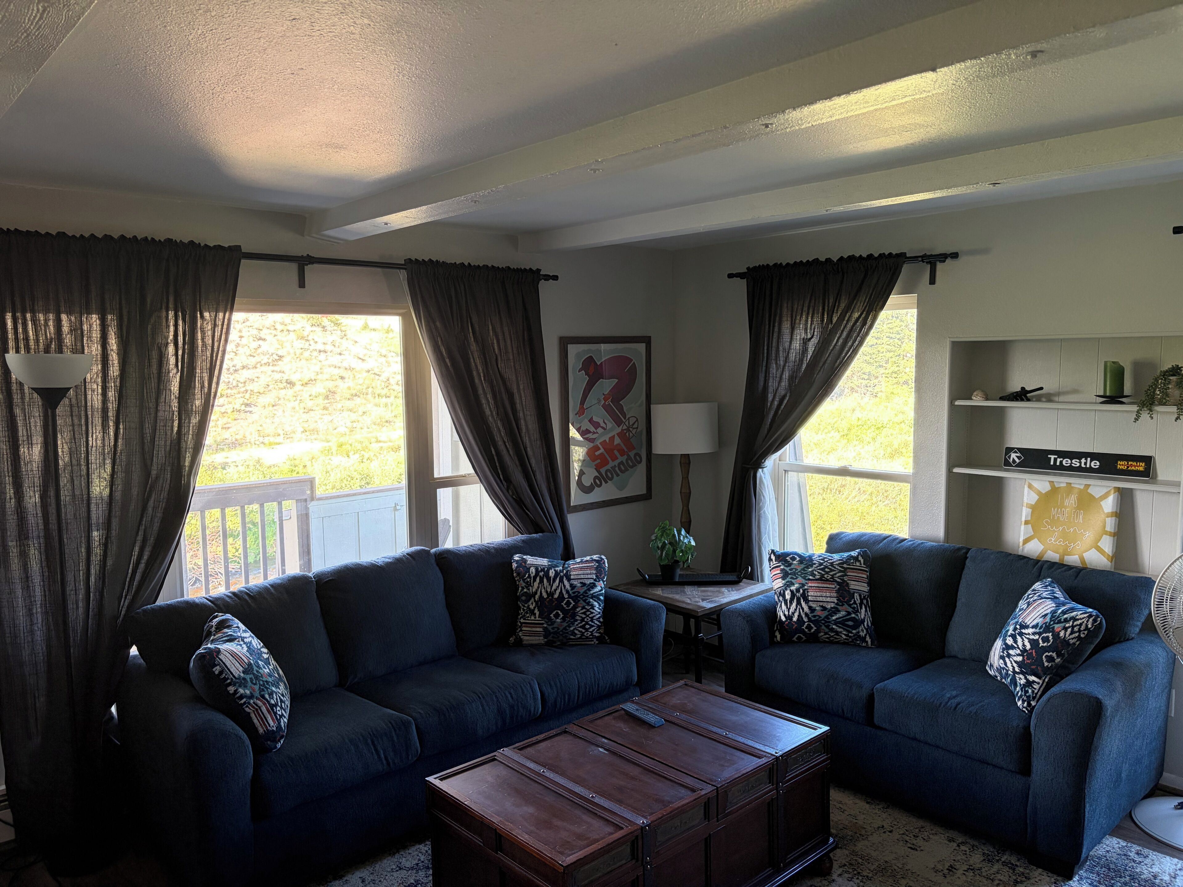 Una sala de estar con dos sofás, una mesa de centro y una lámpara. Se ve una ventana con cortinas, una estantería en la pared con objetos decorativos, y un cuadro enmarcado.