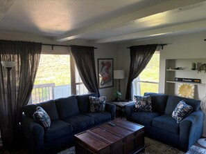 A living room with two sofas, a coffee table, and a lamp. There is a window with curtains, a wall-mounted shelf with decorative items, and a framed picture. - Perfect 3-bedroom condo in lovely Winter Park (Winter Park)